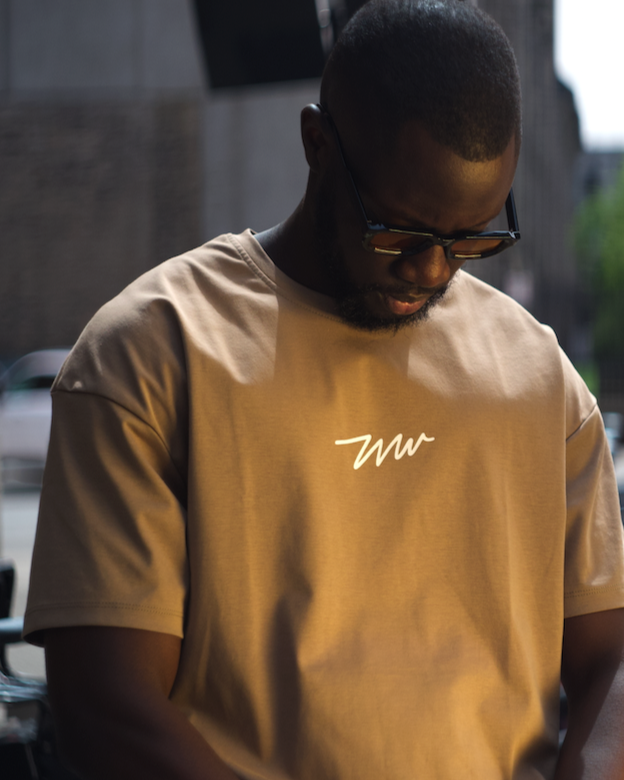Tall man wearing a khaki Wadlow t-shirt, looking down outdoors.