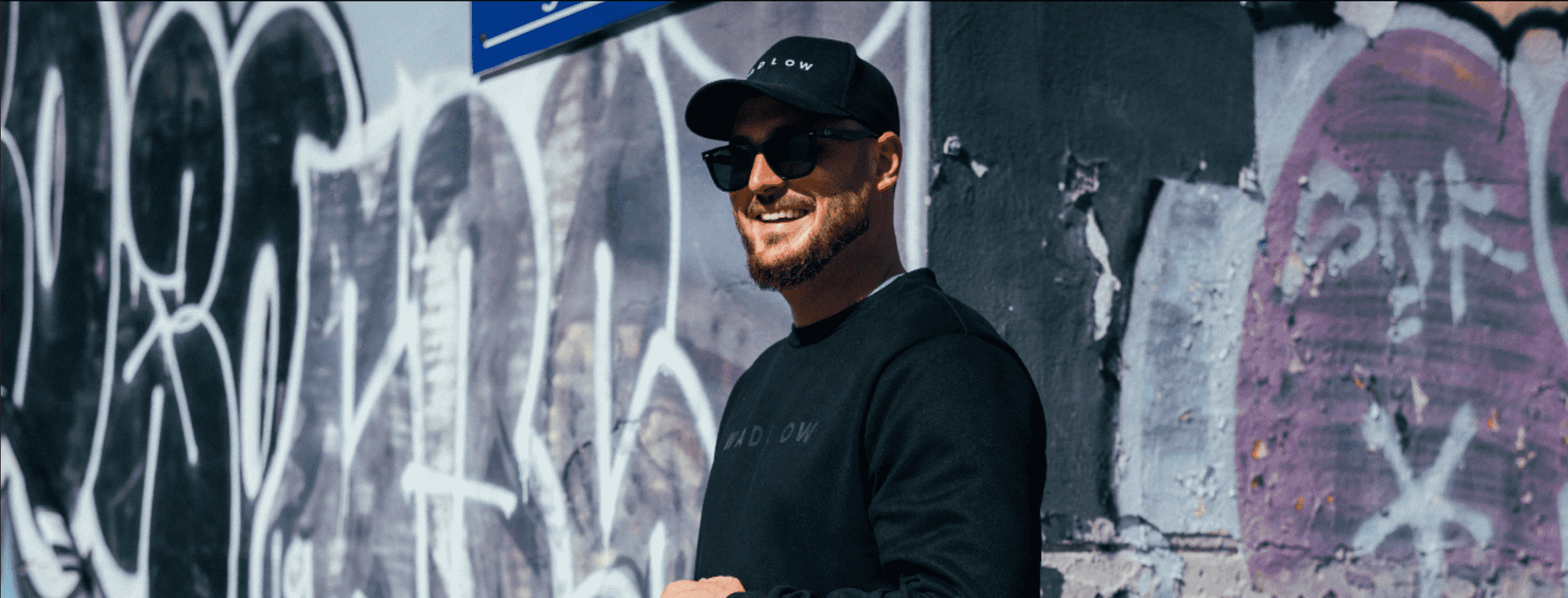a tall Man wearing a black Wadlow crewneck and cap in a graffiti-covered urban setting.